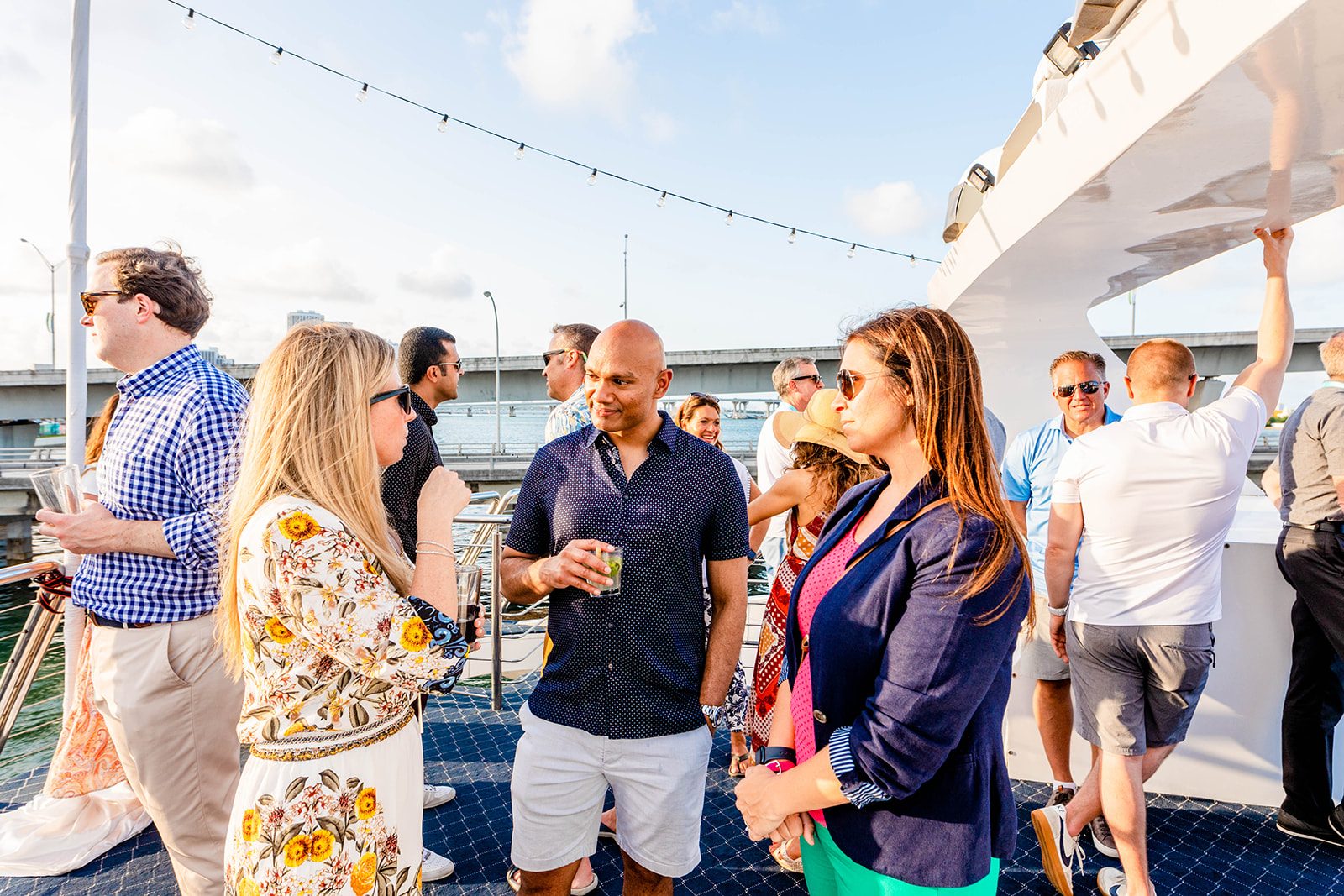 Colleagues talking with cocktails on a yacht deck under string lights at golden hour in Miami