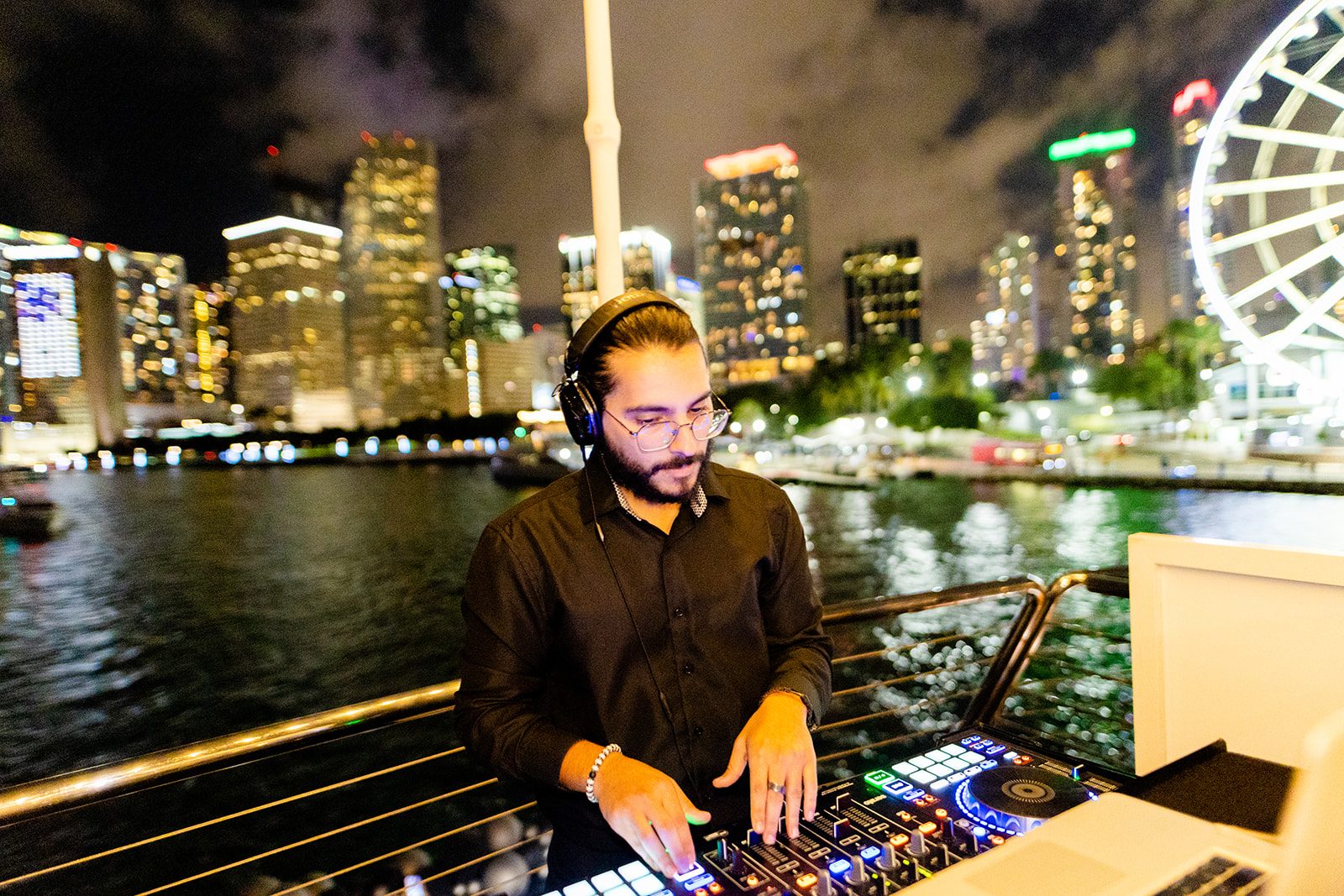 DJ mixing on deck at night with the downtown Miami skyline behind him